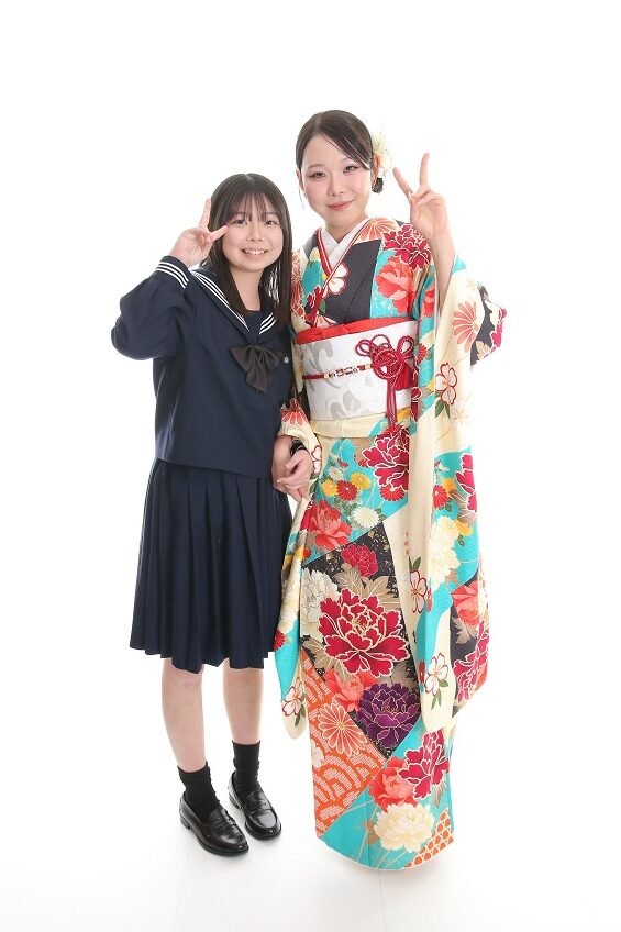 成人式の前撮りでカラフルな振袖を着てピースしている女性とその隣に制服姿の振袖姿の女性の妹がピースしている画像